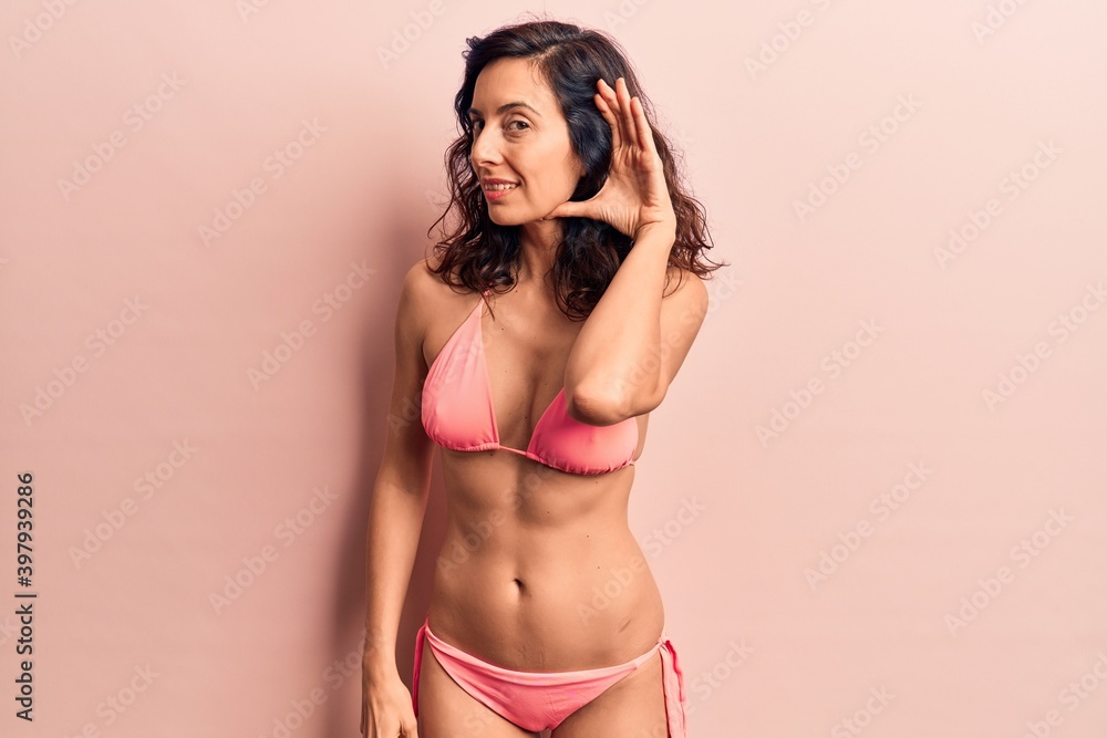 Fototapeta premium Young beautiful hispanic woman wearing bikini smiling with hand over ear listening and hearing to rumor or gossip. deafness concept.