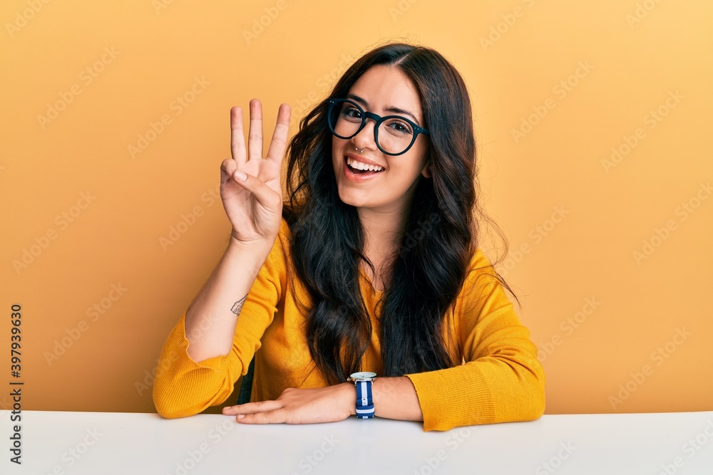 Beautiful brunette young woman wearing glasses and casual clothes sitting on the table showing and pointing up with fingers number three while smiling confident and happy. Beautiful brunette young woman wearing glasses and casual clothes sitting on the table showing and pointing up with fingers number three while smiling confident and happy.