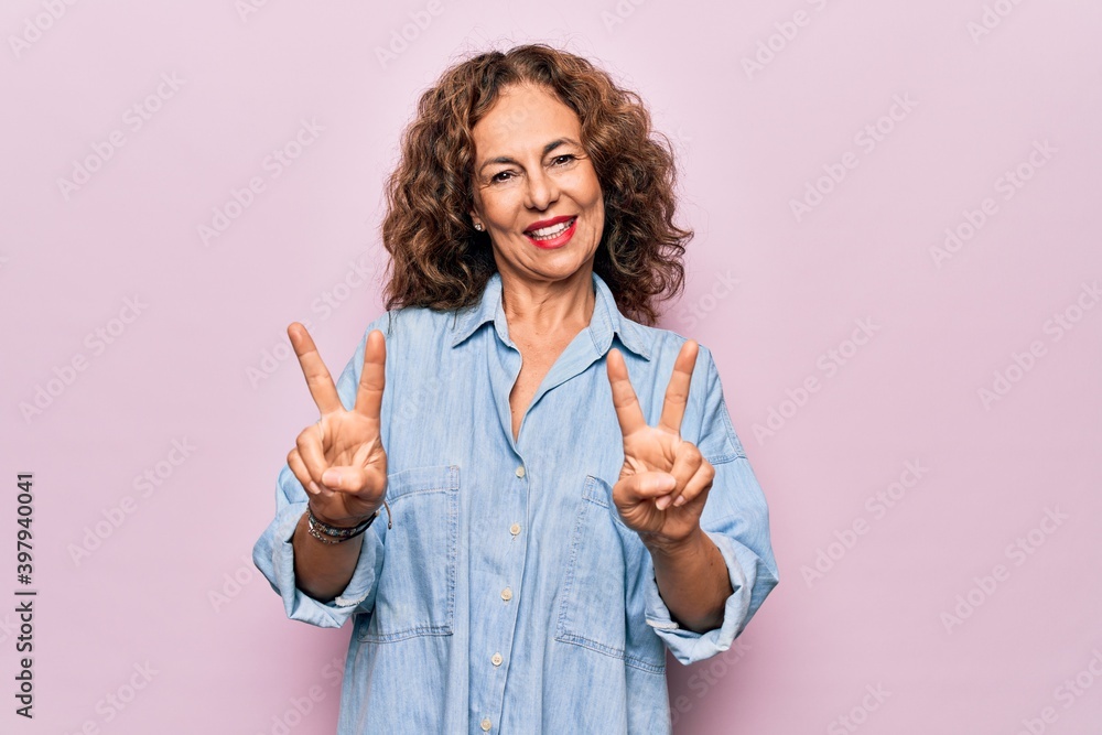 Middle age beautiful woman wearing casual denim shirt standing over pink background smiling with tongue out showing fingers of both hands doing victory sign. Number two. Middle age beautiful woman wearing casual denim shirt standing over pink background smiling with tongue out showing fingers of both hands doing victory sign. Number two.