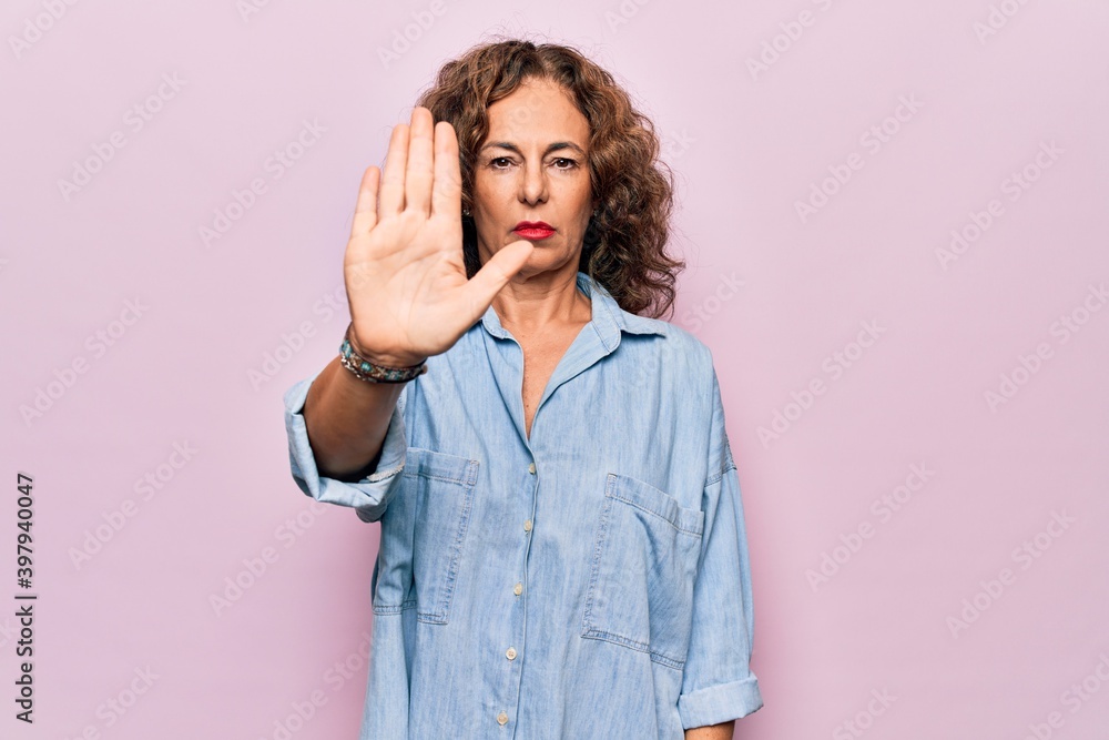 Middle age beautiful woman wearing casual denim shirt standing over pink background doing stop sing with palm of the hand. Warning expression with negative and serious gesture on the face. Middle age beautiful woman wearing casual denim shirt standing over pink background doing stop sing with palm of the hand. Warning expression with negative and serious gesture on the face.