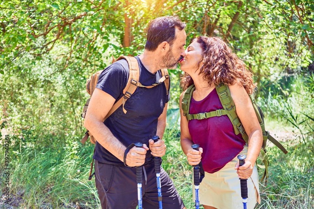 Beautiful couple of hiker wearing backpack kissing. Standing at forest doing trekking using hiking stick over landscape background Beautiful couple of hiker wearing backpack kissing. Standing at forest doing trekking using hiking stick over landscape background