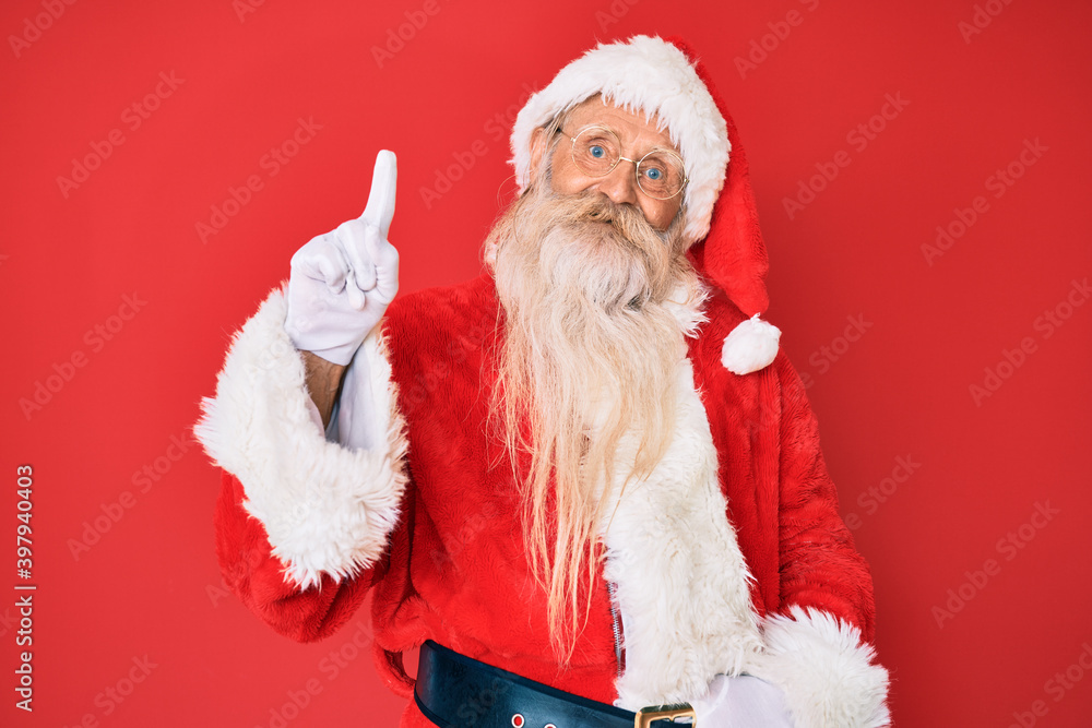 Old senior man with grey hair and long beard wearing traditional santa claus costume showing and pointing up with finger number one while smiling confident and happy.