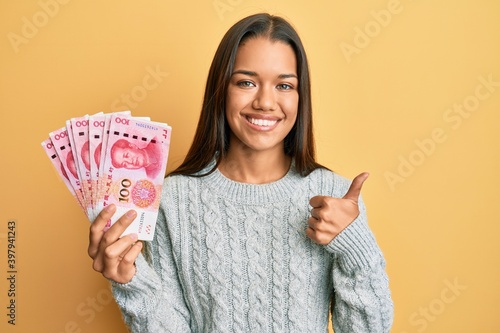 Beautiful hispanic woman holding 100 yuan chinese banknotes smiling happy and positive, thumb up doing excellent and approval sign