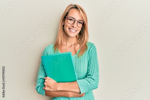 Beautiful blonde woman holding book wearing glasses smiling with a happy and cool smile on face. showing teeth.