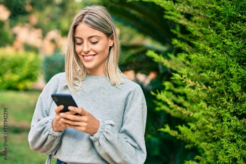 Young blonde girl smiling happy using smartphone at the park.