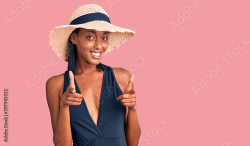 Young african american woman wearing swimsuit and summer hat pointing fingers to camera with happy and funny face. good energy and vibes.
