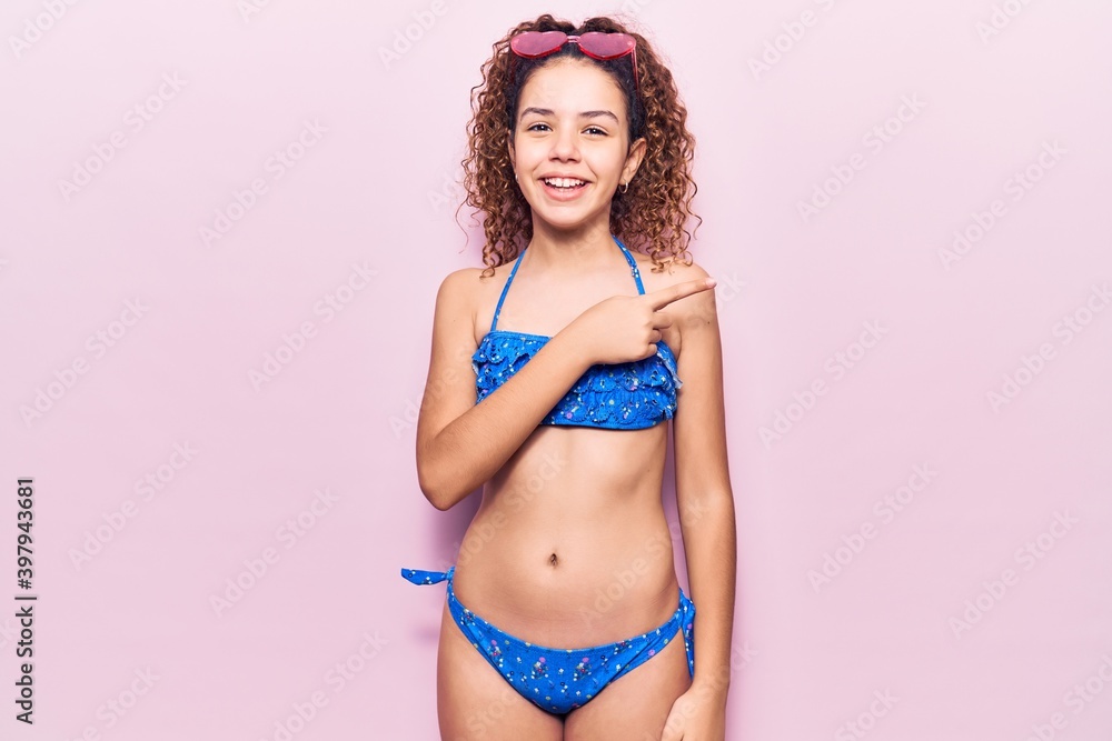 Naklejka premium Beautiful kid girl with curly hair wearing bikini and sunglasses cheerful with a smile of face pointing with hand and finger up to the side with happy and natural expression on face