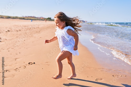 Adorable blonde child wearing summer dress smiling happy. Standing with smile on face playing on the sand at the beach