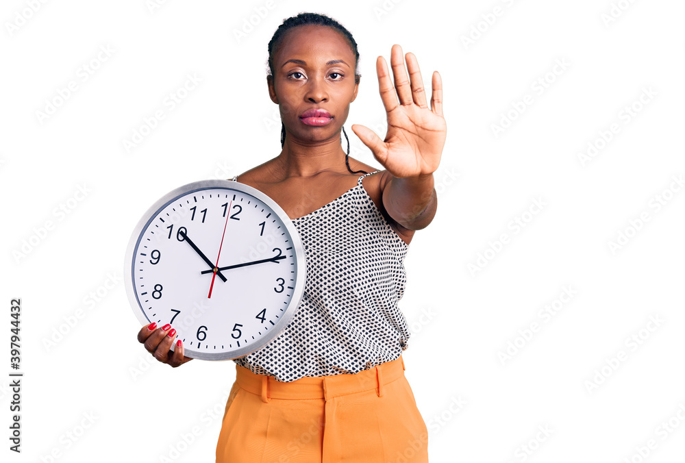 Young african american woman holding big clock with open hand doing stop sign with serious and confident expression, defense gesture Young african american woman holding big clock with open hand doing stop sign with serious and confident expression, defense gesture