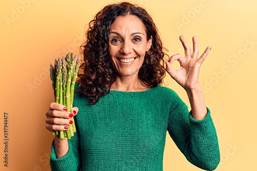 Middle age beautiful woman holding asparagus doing ok sign with fingers, smiling friendly gesturing excellent symbol