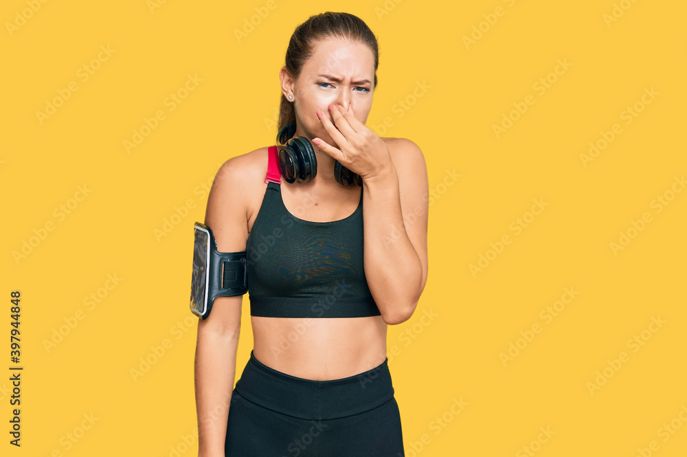 Beautiful Blonde Woman Wearing Gym Clothes And Using Headphones Smelling Something Stinky And