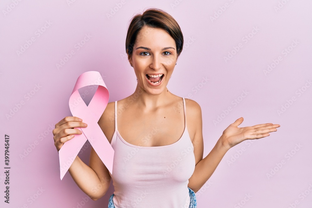 Young brunette woman with short hair holding pink cancer ribbon celebrating achievement with happy smile and winner expression with raised hand Young brunette woman with short hair holding pink cancer ribbon celebrating achievement with happy smile and winner expression with raised hand