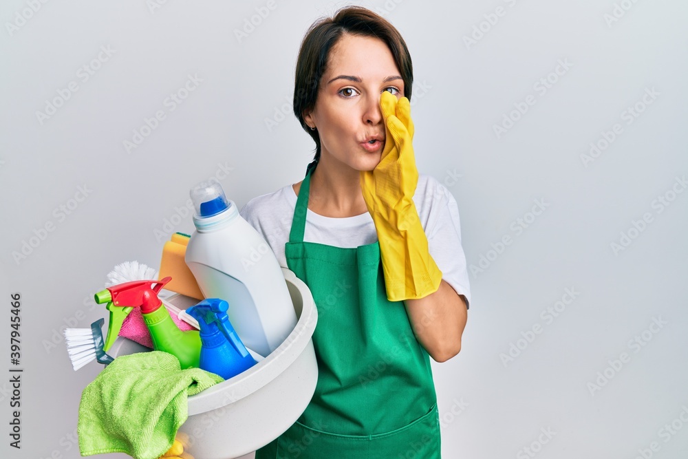 Young brunette woman with short hair wearing apron holding cleaning products hand on mouth telling secret rumor, whispering malicious talk conversation Young brunette woman with short hair wearing apron holding cleaning products hand on mouth telling secret rumor, whispering malicious talk conversation