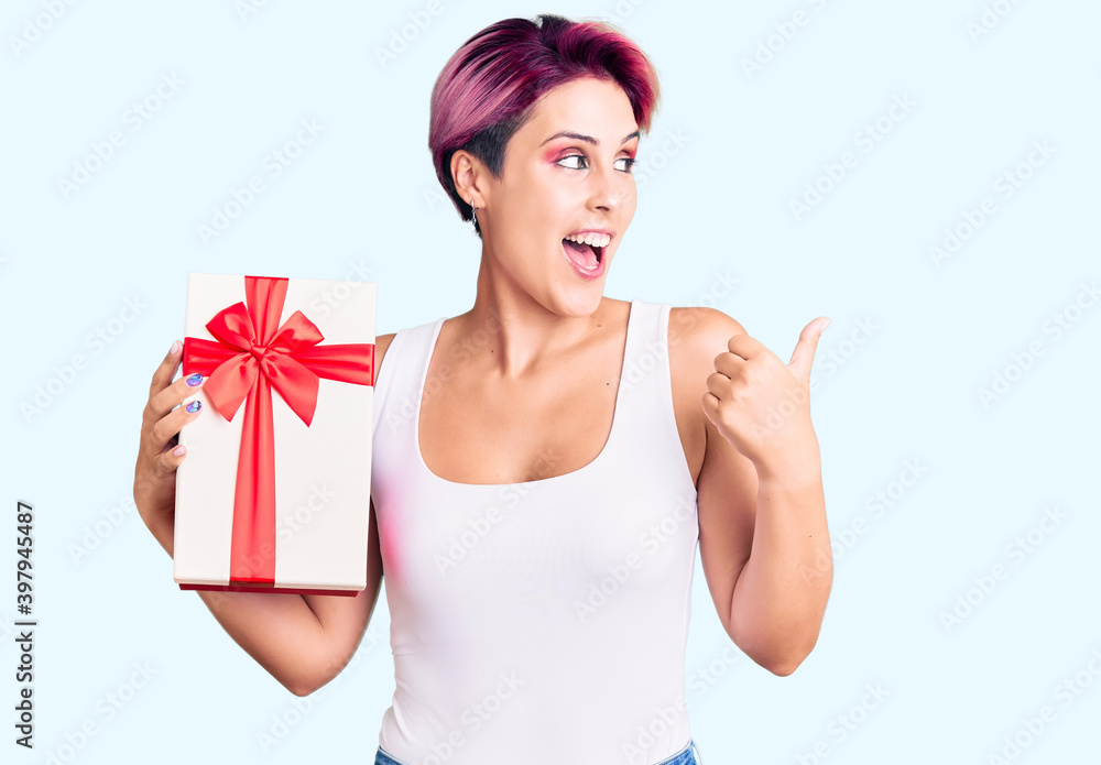 Young beautiful woman with pink hair holding gift pointing thumb up to the side smiling happy with open mouth Young beautiful woman with pink hair holding gift pointing thumb up to the side smiling happy with open mouth