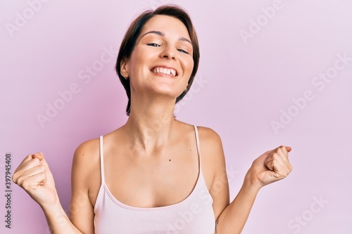 Young brunette woman with short hair over pink background very happy and excited doing winner gesture with arms raised, smiling and screaming for success. celebration concept.
