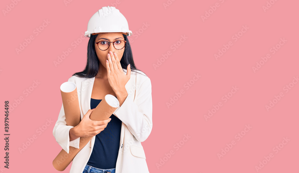 Young beautiful latin girl wearing architect hardhat holding blueprints covering mouth with hand, shocked and afraid for mistake. surprised expression Young beautiful latin girl wearing architect hardhat holding blueprints covering mouth with hand, shocked and afraid for mistake. surprised expression