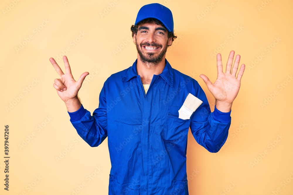 Handsome young man with curly hair and bear wearing builder jumpsuit uniform showing and pointing up with fingers number eight while smiling confident and happy. Handsome young man with curly hair and bear wearing builder jumpsuit uniform showing and pointing up with fingers number eight while smiling confident and happy.