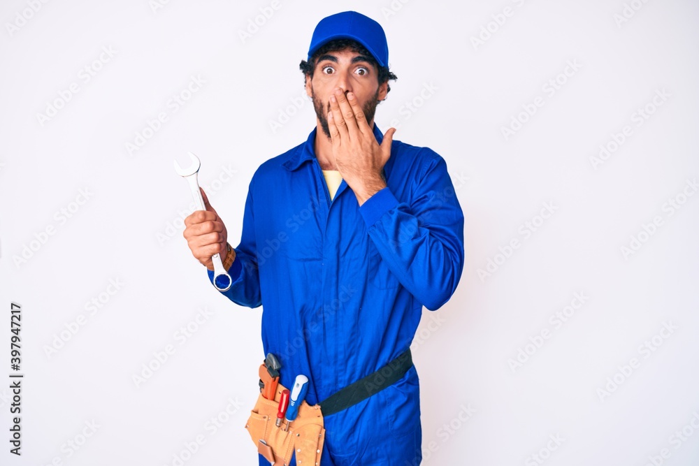 Handsome young man with curly hair and bear wearing builder jumpsuit uniform and holding wrench covering mouth with hand, shocked and afraid for mistake. surprised expression