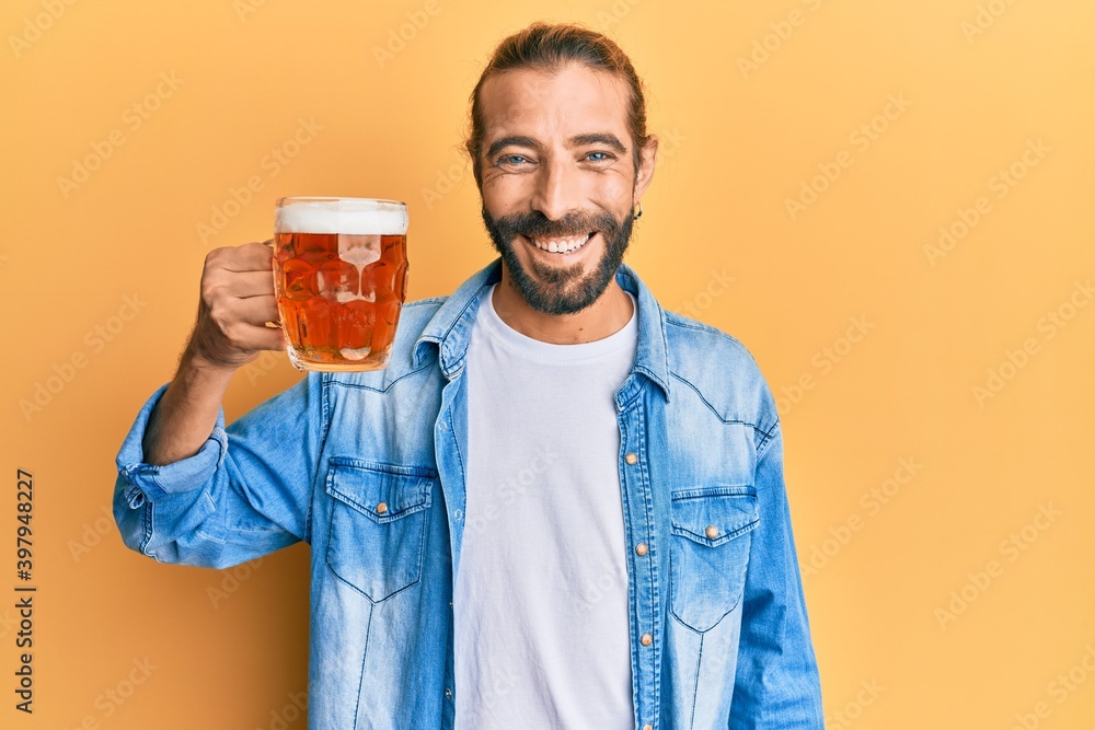 Attractive man with long hair and beard drinking a pint of beer looking positive and happy standing and smiling with a confident smile showing teeth Attractive man with long hair and beard drinking a pint of beer looking positive and happy standing and smiling with a confident smile showing teeth