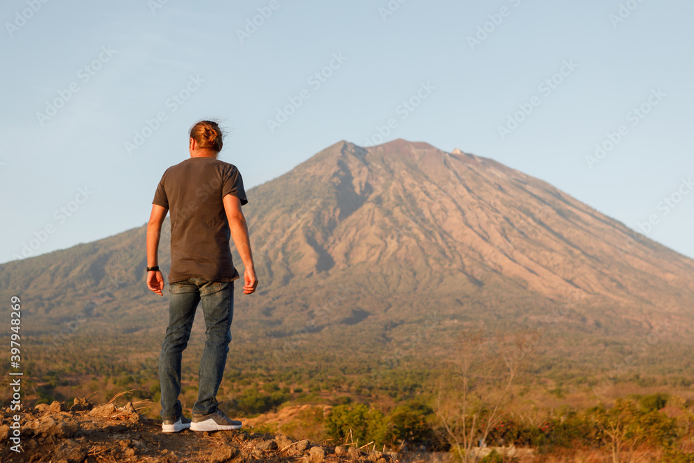 Rear view young man traveler standing on top of rock and enjoying amazing volcanic mountain Agung view in Bali during sunrise. Travel and active lifestyle concept. Rear view young man traveler standing on top of rock and enjoying amazing volcanic mountain Agung view in Bali during sunrise. Travel and active lifestyle concept.