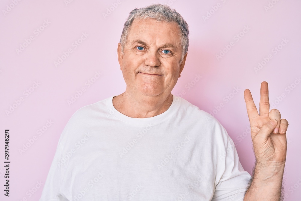 Senior grey-haired man wearing casual white tshirt smiling with happy face winking at the camera doing victory sign. number two. Senior grey-haired man wearing casual white tshirt smiling with happy face winking at the camera doing victory sign. number two.