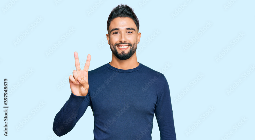 Young man with beard wearing casual blue winter sweater smiling with happy face winking at the camera doing victory sign. number two. Young man with beard wearing casual blue winter sweater smiling with happy face winking at the camera doing victory sign. number two.