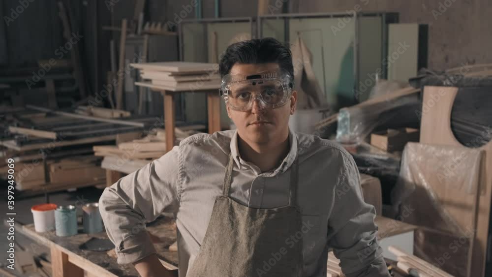 Medium close-up portrait of man working as carpenter in small joinery looking at camera wearing safety glasses for woodworking with piles of wood planks in background Medium close-up portrait of man working as carpenter in small joinery looking at camera wearing safety glasses for woodworking with piles of wood planks in background