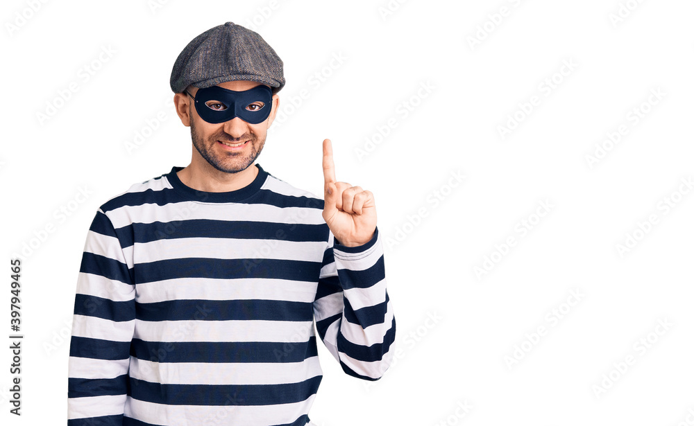 Young handsome man wearing burglar mask showing and pointing up with finger number one while smiling confident and happy. Young handsome man wearing burglar mask showing and pointing up with finger number one while smiling confident and happy.