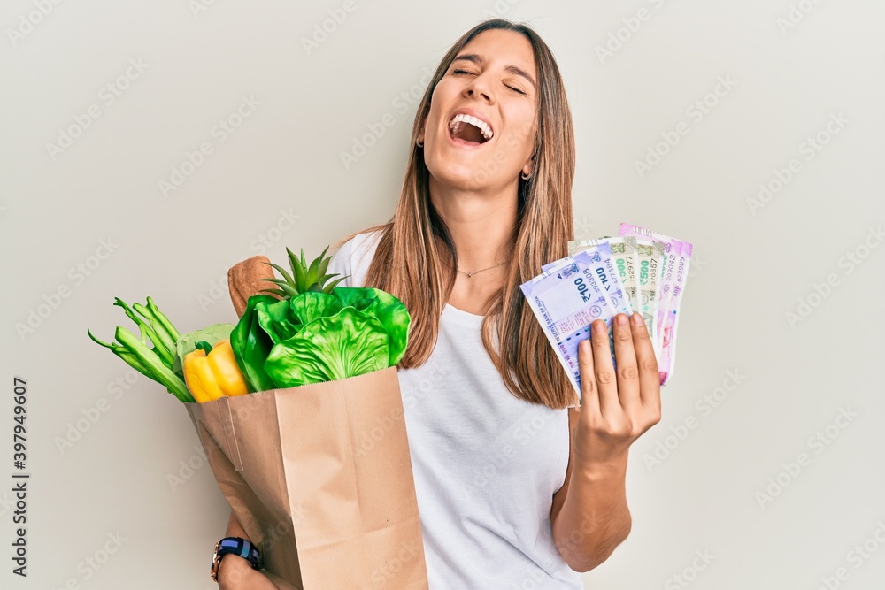 Brunette young woman holding groceries and indian rupee banknotes smiling and laughing hard out loud because funny crazy joke. Brunette young woman holding groceries and indian rupee banknotes smiling and laughing hard out loud because funny crazy joke.