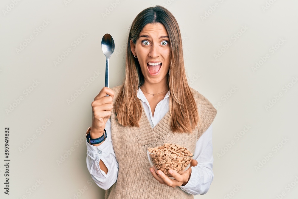 Brunette young woman eating healthy whole grain cereals with spoon celebrating crazy and amazed for success with open eyes screaming excited. Brunette young woman eating healthy whole grain cereals with spoon celebrating crazy and amazed for success with open eyes screaming excited.