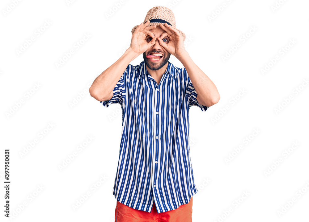 Young handsome man with beard wearing summer hat and striped shirt doing ok gesture like binoculars sticking tongue out, eyes looking through fingers. crazy expression. Young handsome man with beard wearing summer hat and striped shirt doing ok gesture like binoculars sticking tongue out, eyes looking through fingers. crazy expression.
