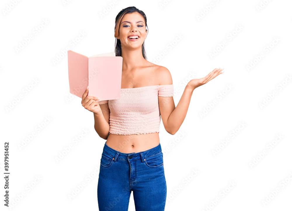 Young beautiful woman holding book celebrating victory with happy smile and winner expression with raised hands Young beautiful woman holding book celebrating victory with happy smile and winner expression with raised hands