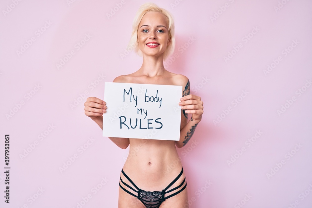 Young blonde woman with tattoo standing shirtless holding banner with my body my rules message smiling with a happy and cool smile on face. showing teeth. Young blonde woman with tattoo standing shirtless holding banner with my body my rules message smiling with a happy and cool smile on face. showing teeth.