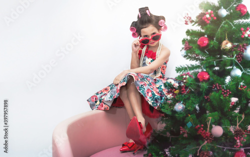 Stylish little girl portrait has curlers in hair wearing beautiful dress and red mom's shoes, sit near the Christmas tree