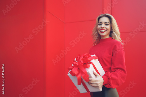 Happy blonde woman in holding gift box and looking at camera while enjoys over red background. High quality photo.