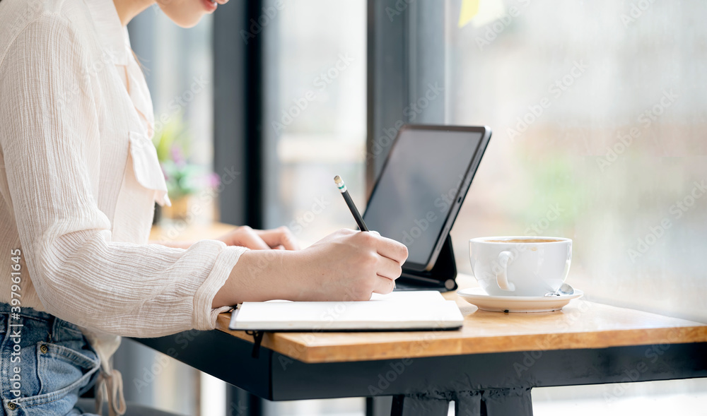 Shot of female hand writing on notebook with pencil while sitting at the table with laptop and cup of coffee. Shot of female hand writing on notebook with pencil while sitting at the table with laptop and cup of coffee.