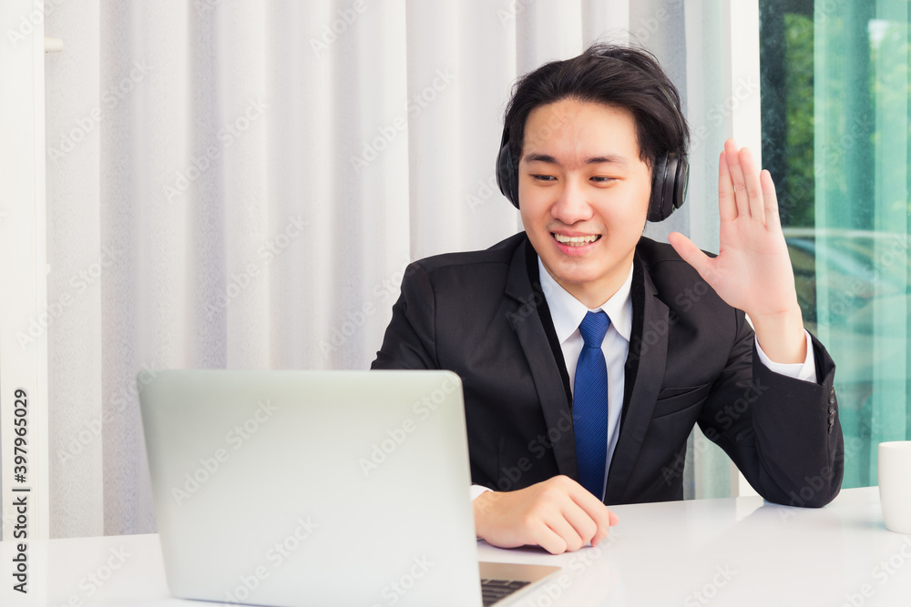 Work from home, Asian young businessman smile wearing headphones and suit video conference call or facetime on desk and raise your hand to say hello greet colleague at home office Work from home, Asian young businessman smile wearing headphones and suit video conference call or facetime on desk and raise your hand to say hello greet colleague at home office