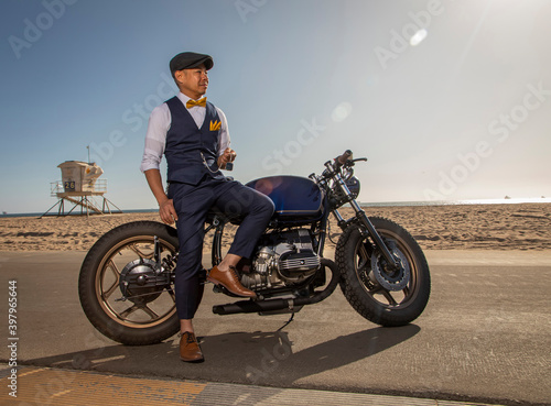 An Asian man dressed in vintage clothing sitting on a motorcycle at the beach