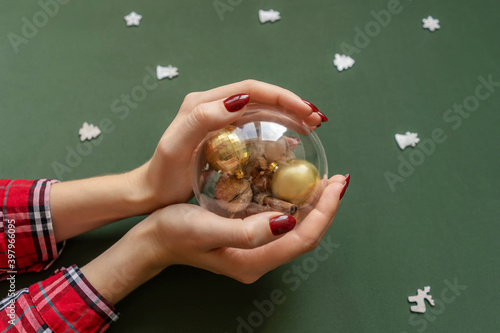 Christmas manicure. Red nails, hand with transparent ball with christmas knick-knackery inside on green background with silver baubles around.