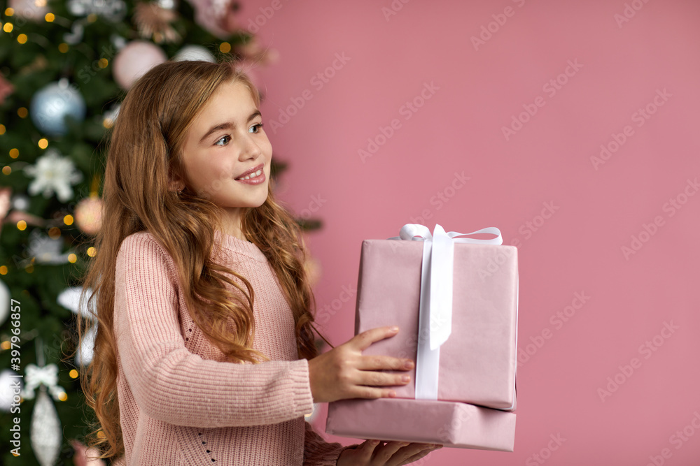 cute smiling little child girl holding Christmas gift box in hand over the pink background and christmas tree. copy space cute smiling little child girl holding Christmas gift box in hand over the pink background and christmas tree. copy space