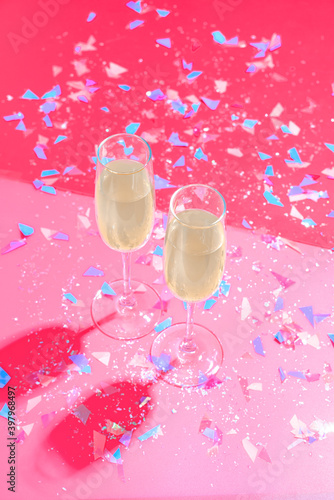 Two glasses of champagne stand on a double pink background strewn with glittering holographic confetti