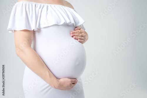 Motherhood, pregnancy, expecting a baby, family concept. A pregnant woman in a beautiful white tight-fitting dress gently holds her hands on her stomach. Close-up. Side view. Studio white background.