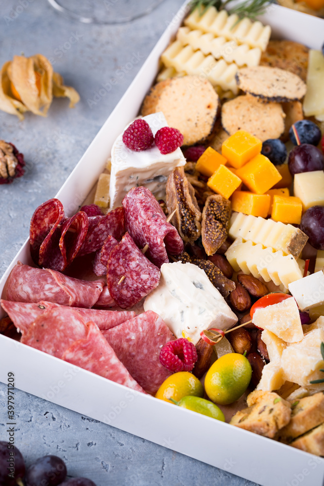 Cheese plate with delicious snacks. Christmas gift box with assorted cheese cubes, meat slices, fruits and nuts in gift box with crackers, olives and wine glass. Delicious charcuterie, copy space