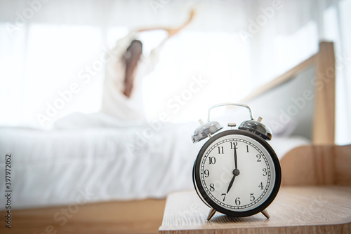 Closeup alarm clock having a good day with background happy woman stretching in bed after waking up, sunlight in morning.