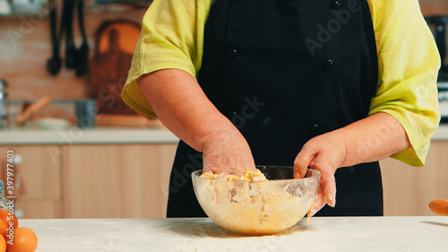 Woman preparing tasty food using wheat flour and cracked fresh eggs in home. Close up of retired elderly chef with bonete kneading in glass bowl pastry ingredients baking homemade cake and bread.