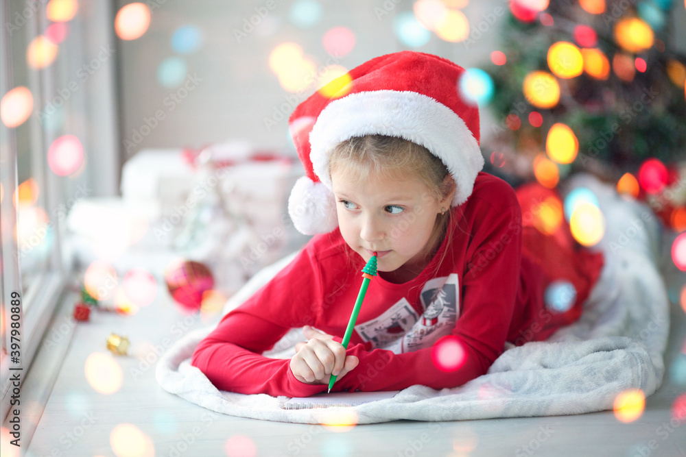 A smiling girl in a Santa hat writes a letter, dreams of gifts to Santa Claus. Child lying on a blanket on the floor of a house with christmas lights A smiling girl in a Santa hat writes a letter, dreams of gifts to Santa Claus. Child lying on a blanket on the floor of a house with christmas lights