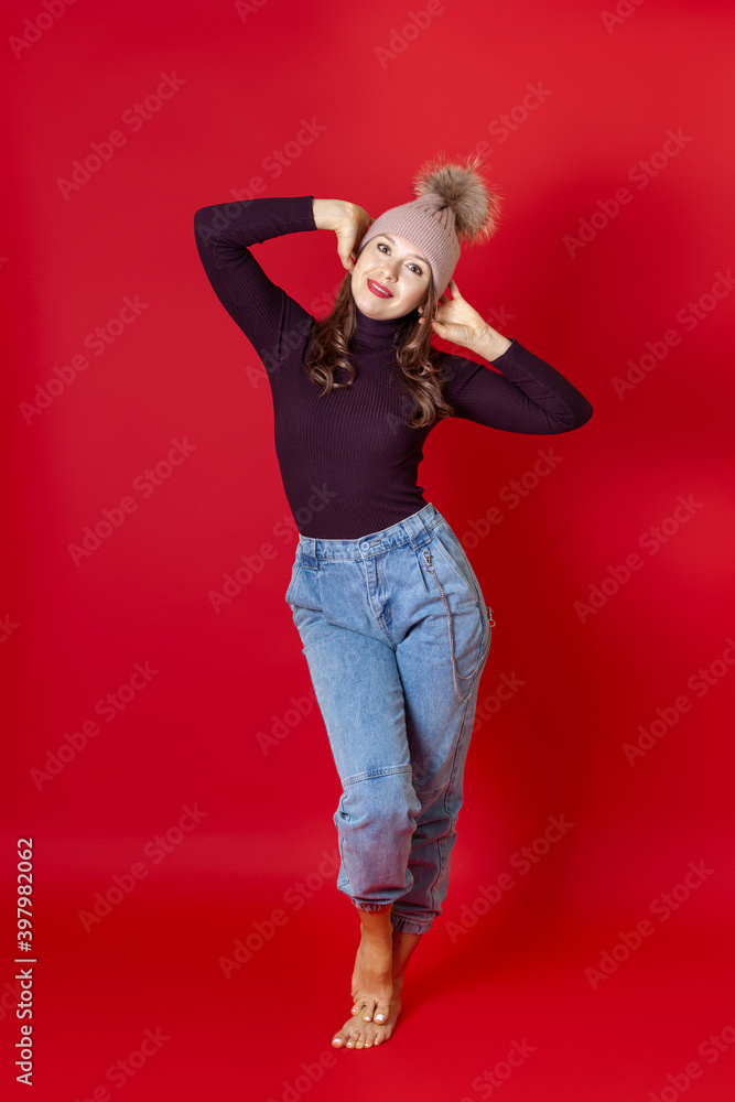 a full-length smiling young woman stands in a knitted winter hat and holds her head with her hands, isolated on a red background. a full-length smiling young woman stands in a knitted winter hat and holds her head with her hands, isolated on a red background.