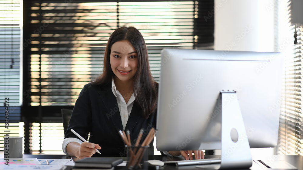Young businesswoman smiling to camera while making notes in notebook at her workspace. Young businesswoman smiling to camera while making notes in notebook at her workspace.