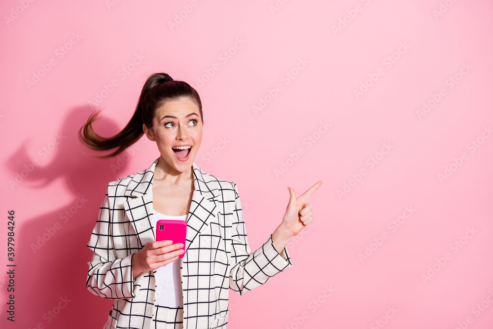 Photo portrait of excited woman pointing finger at blank space holding phone in one hand isolated on pastel pink colored background Photo portrait of excited woman pointing finger at blank space holding phone in one hand isolated on pastel pink colored background