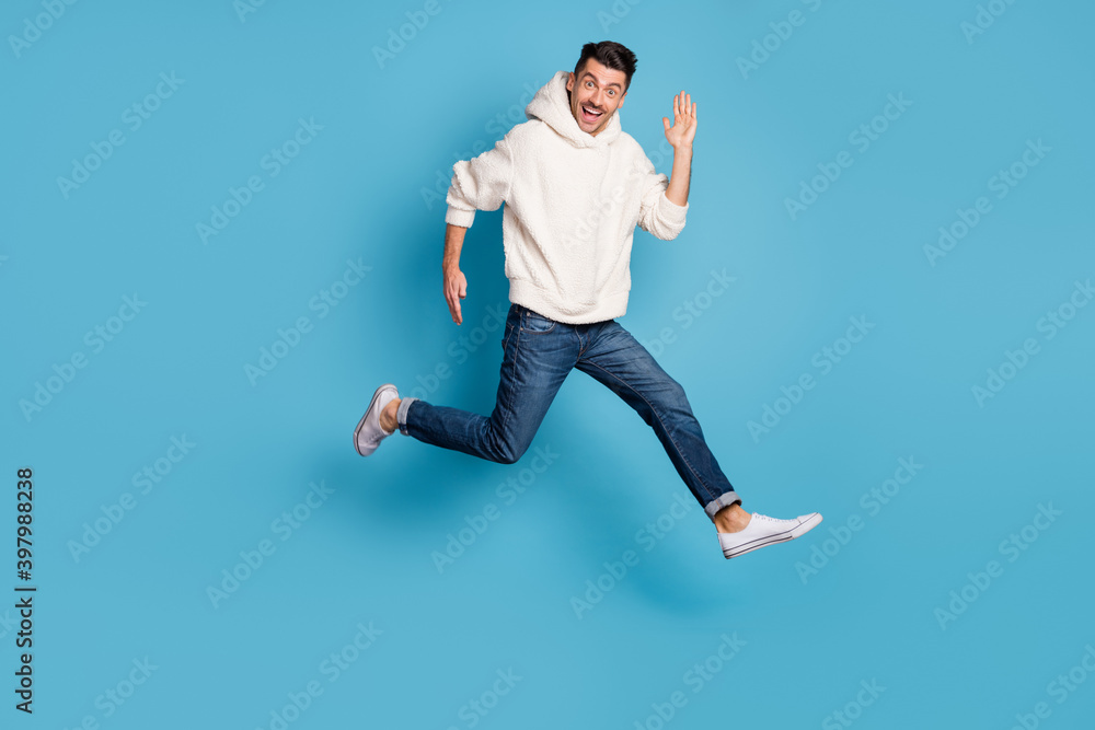Full size photo of young excited positive happy smiling man jumping running say hi isolated on blue color background Full size photo of young excited positive happy smiling man jumping running say hi isolated on blue color background